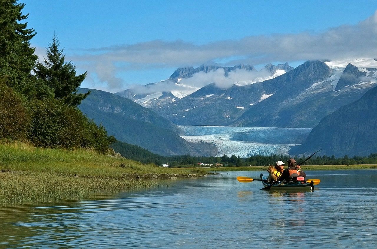 Anglers in a kayak on a calm Alaska lake with a glacier and snowy peaks behind