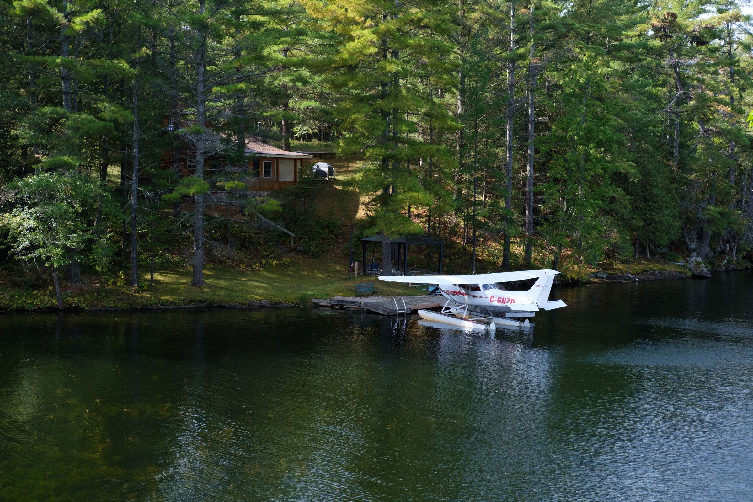 A floatplane moored at a dock below a cabin in tall pines on a quiet lake