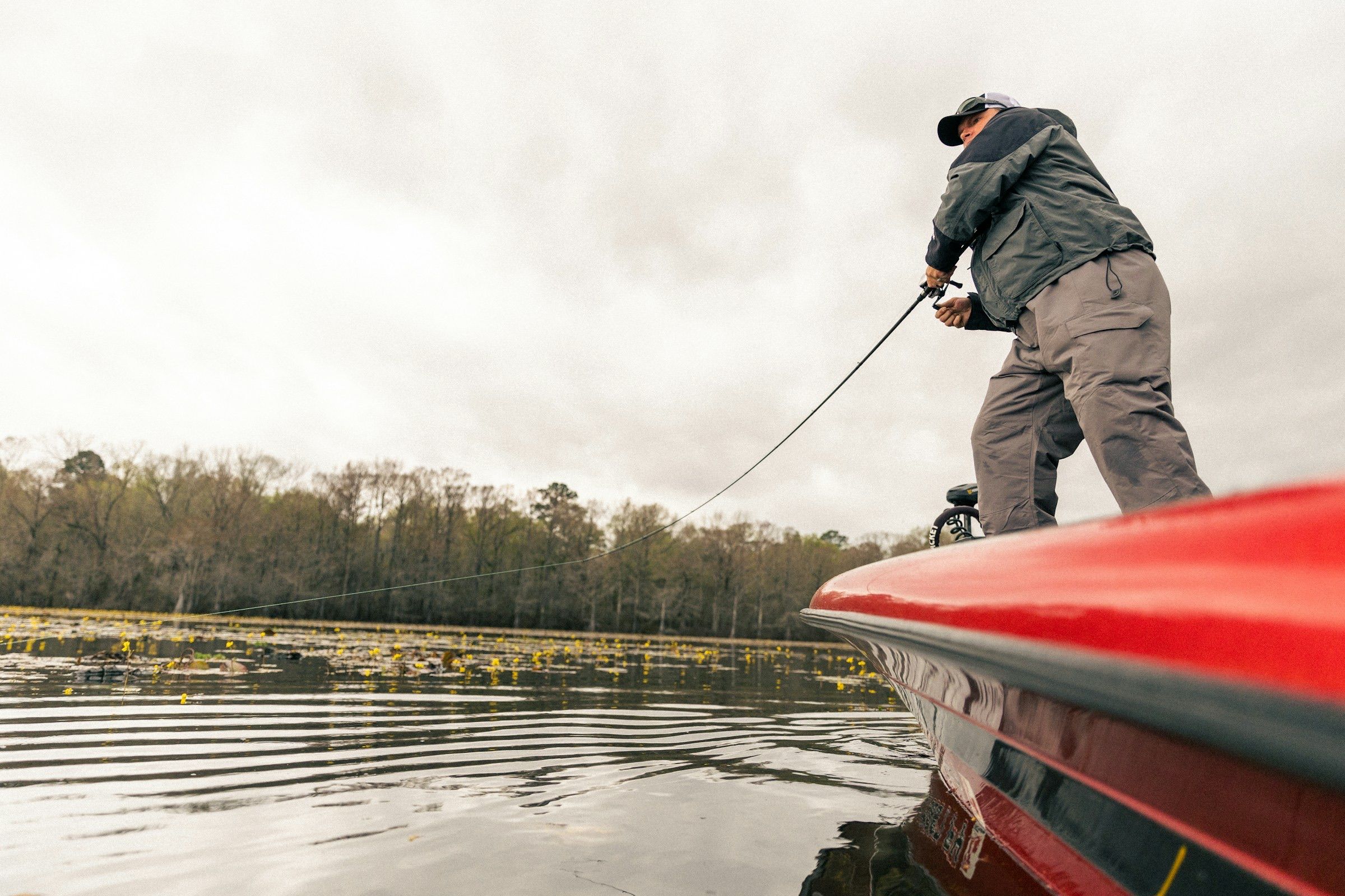 Angler on the bow of a red bass boat reeling in on a lake lined with bare early-spring trees