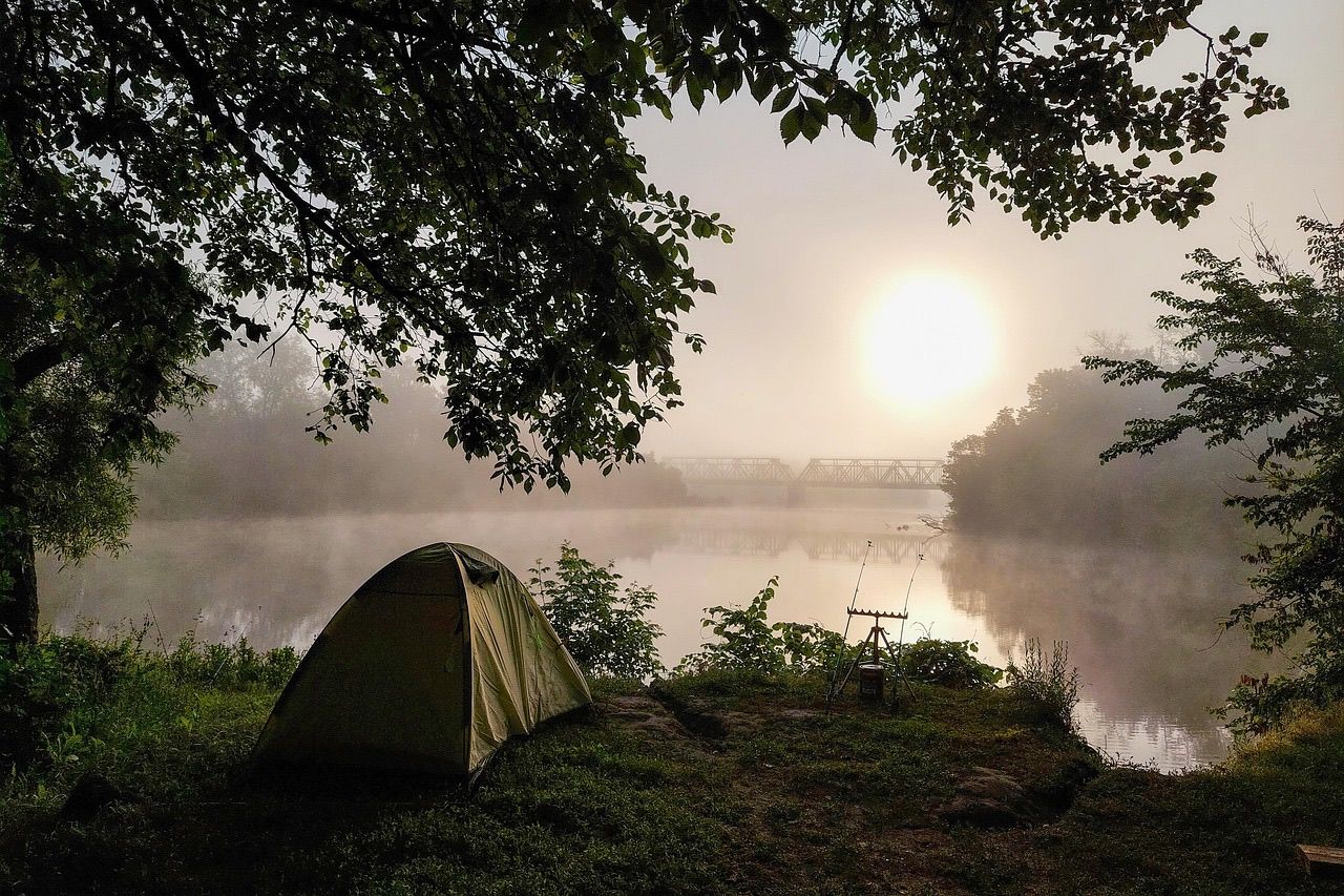 A small tent and two fishing rods on a stand at a riverside campsite as the sun rises through morning mist