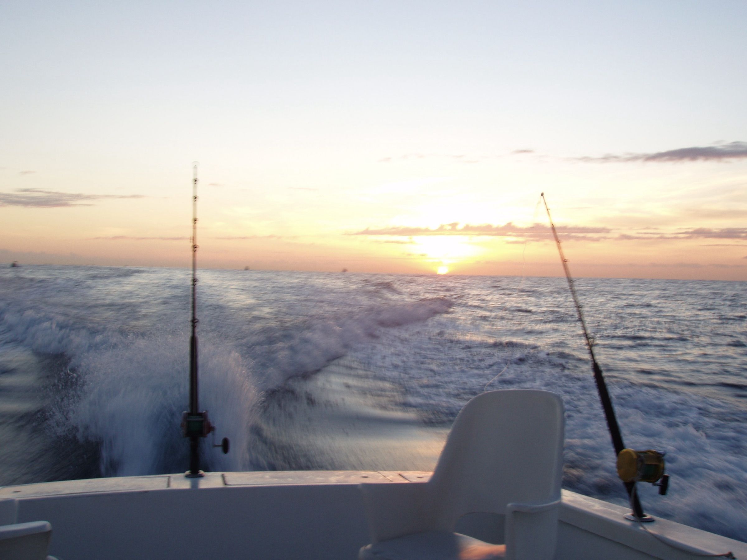 Trolling rods set in the stern rod holders of a fishing boat running offshore at sunrise, with white wake behind
