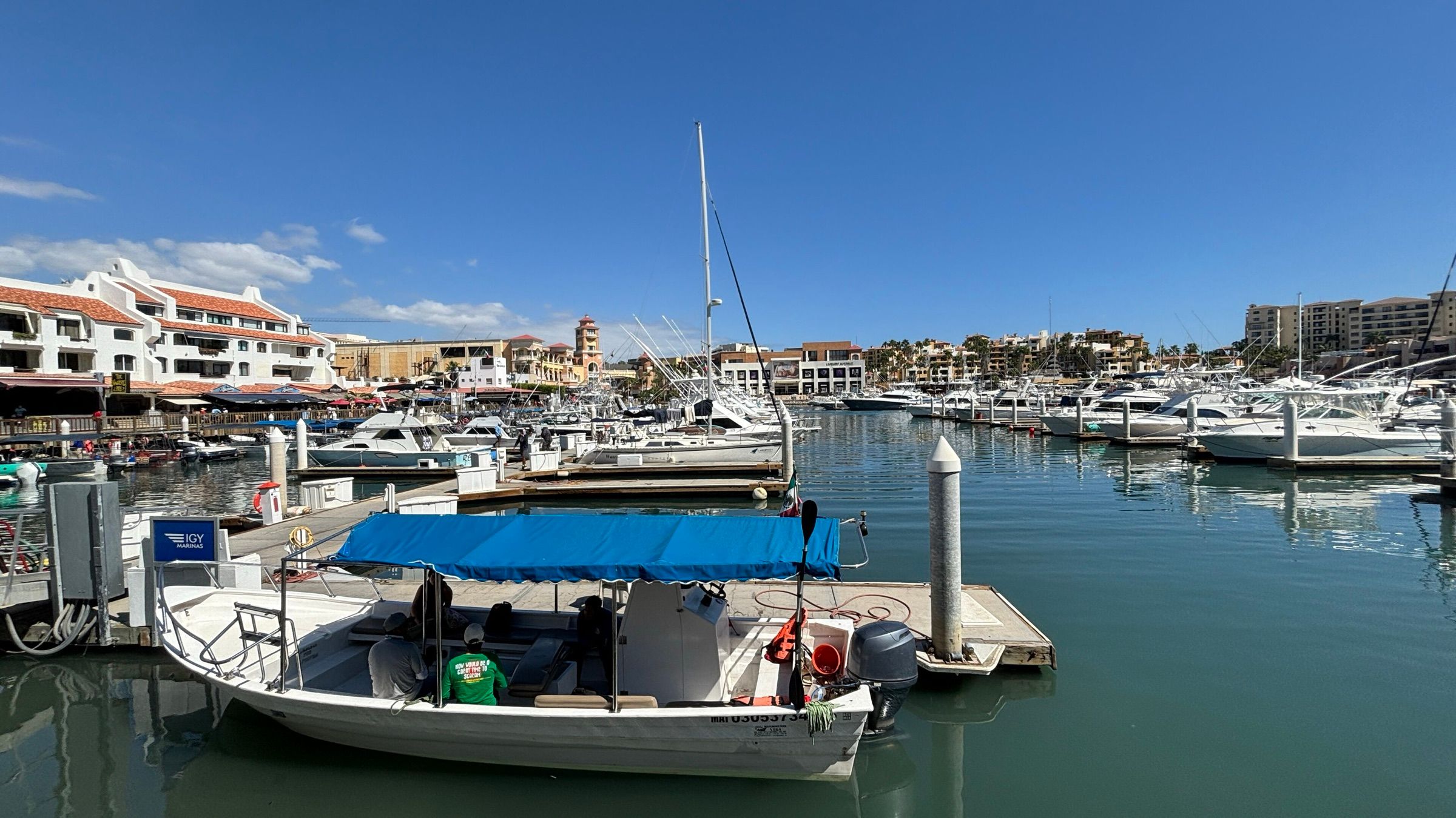 A small open fishing boat with a blue canopy tied up in front of larger yachts and sportfishing boats in a Cabo San Lucas marina