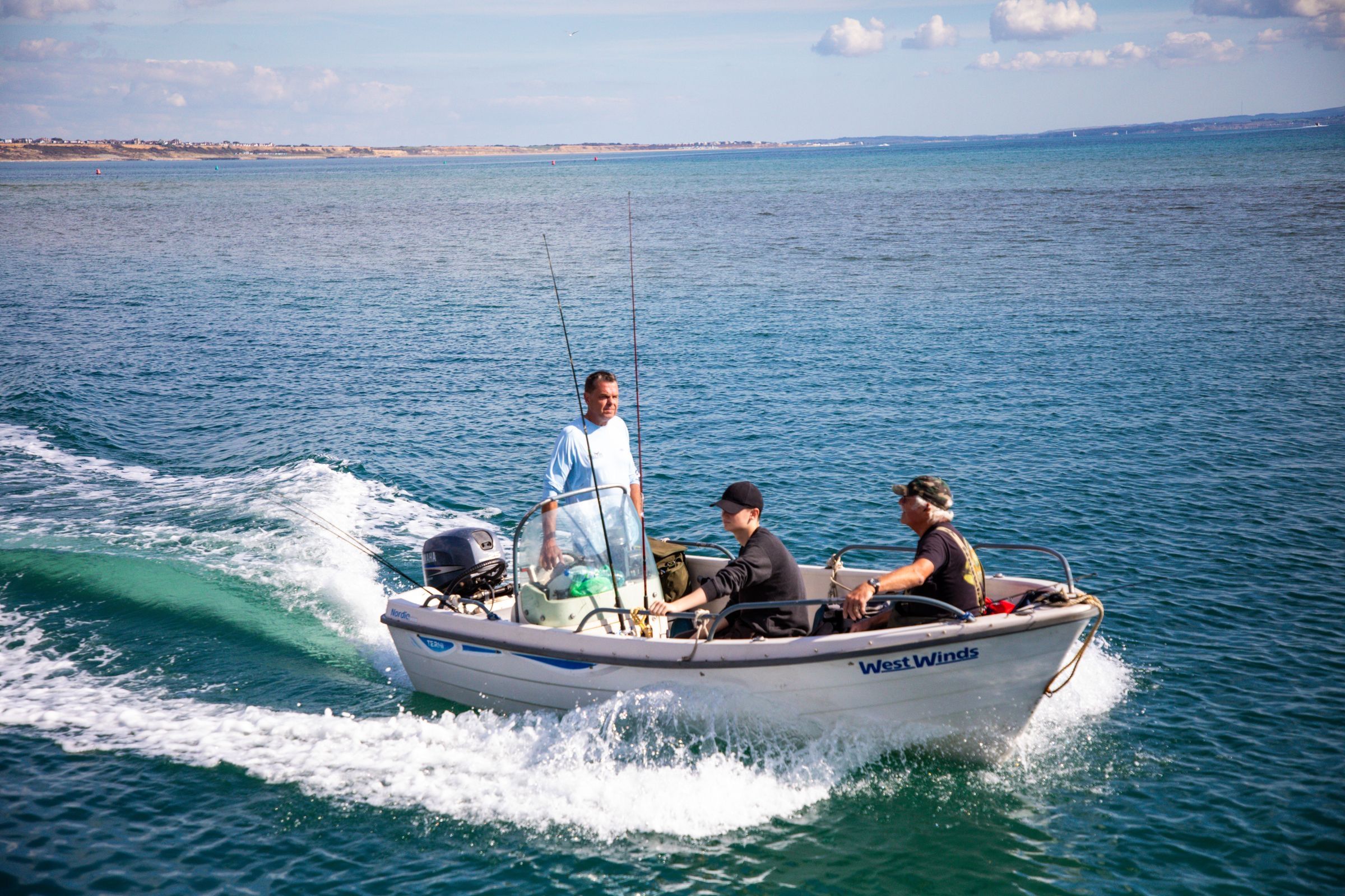 A skipper stands at the console of a small open fishing boat with rods aboard as two anglers ride along on open water off England