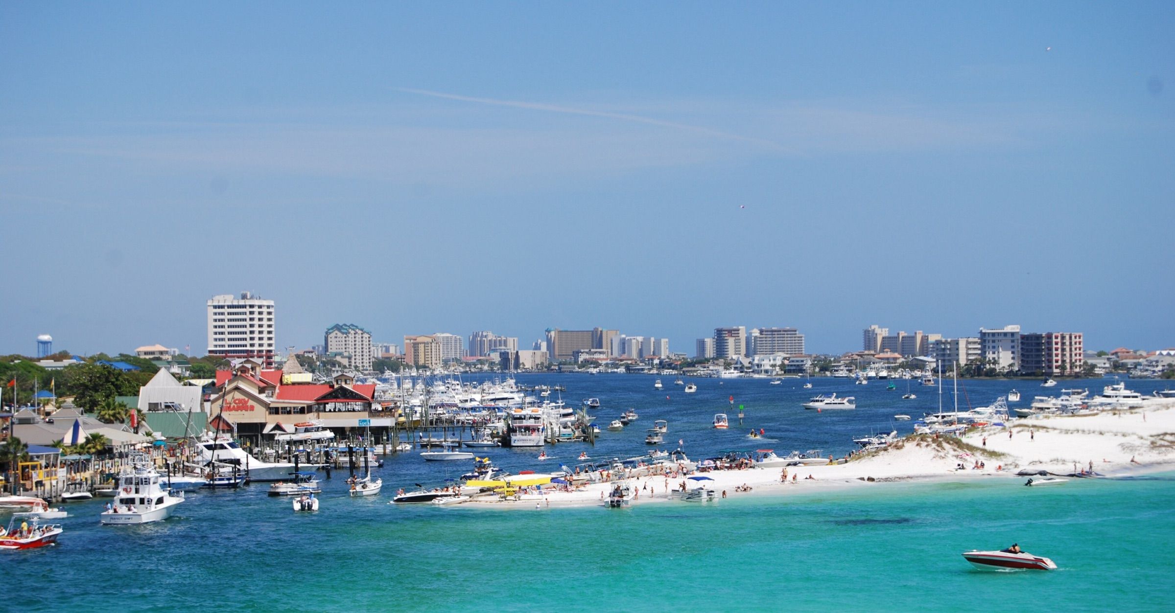 Destin Harbor packed with charter and pleasure boats beside a white-sand point and emerald water, with high-rise condos beyond