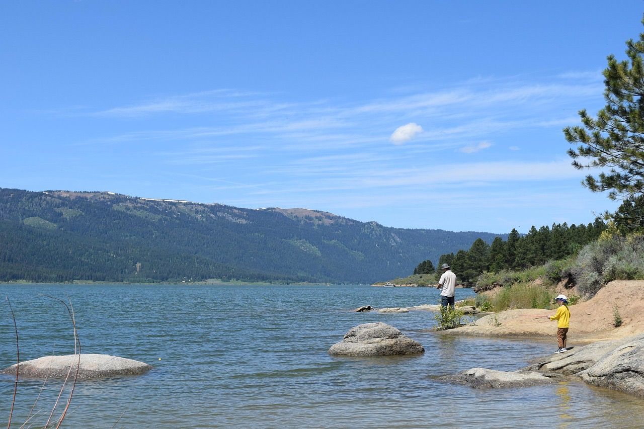 An adult and a small child in a yellow jacket fishing from the rocky shore of a mountain lake on a sunny day