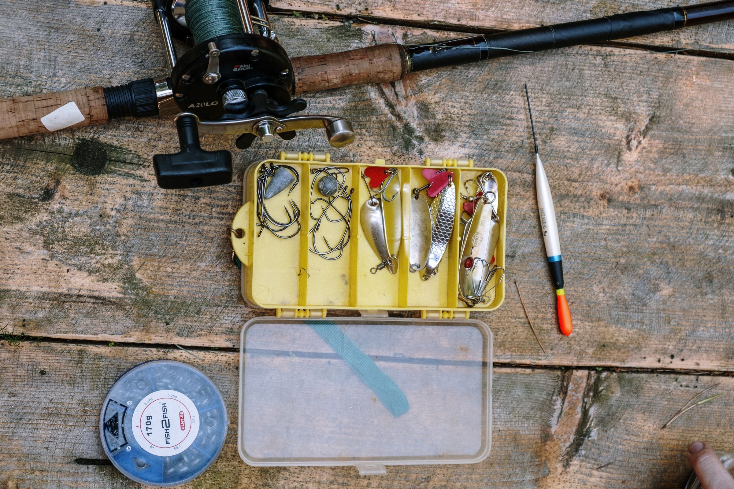 Top view of a rod and reel, a tray of spoons and hooks, a spool of line and a float laid out on weathered boards