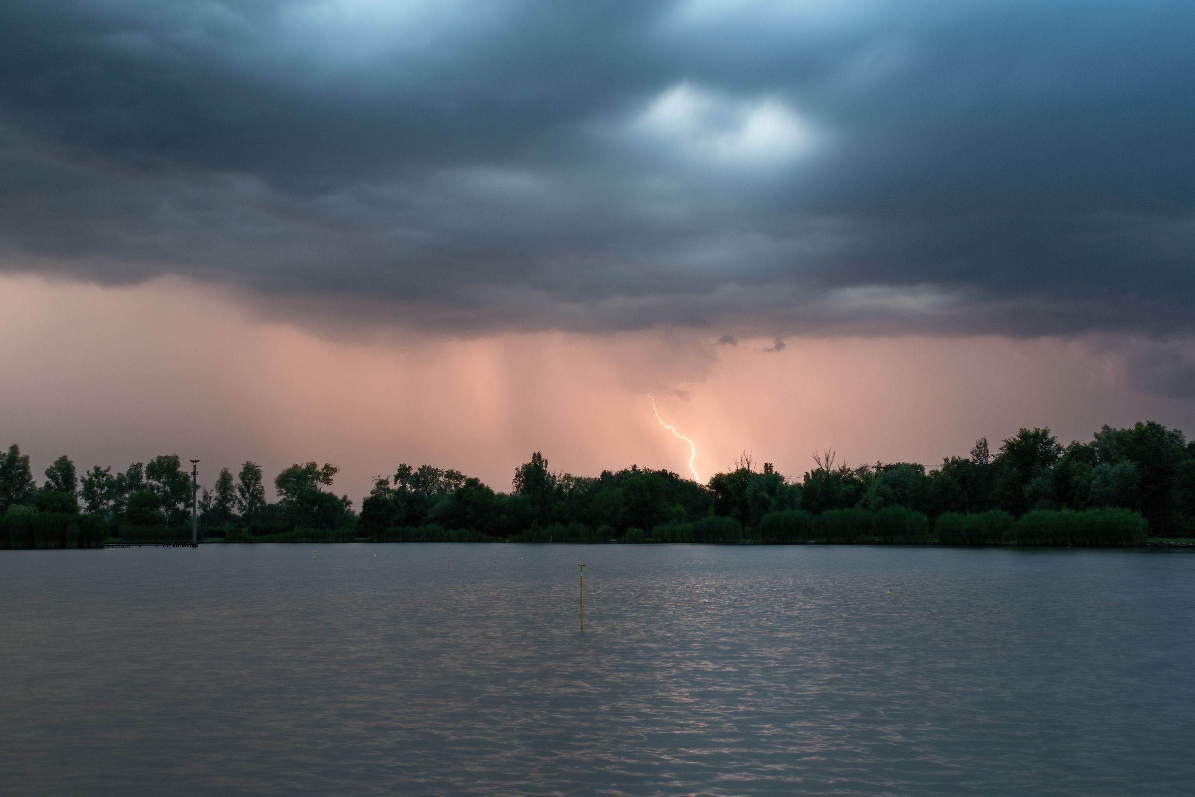 Lightning strikes under dark storm clouds over a calm lake and tree line