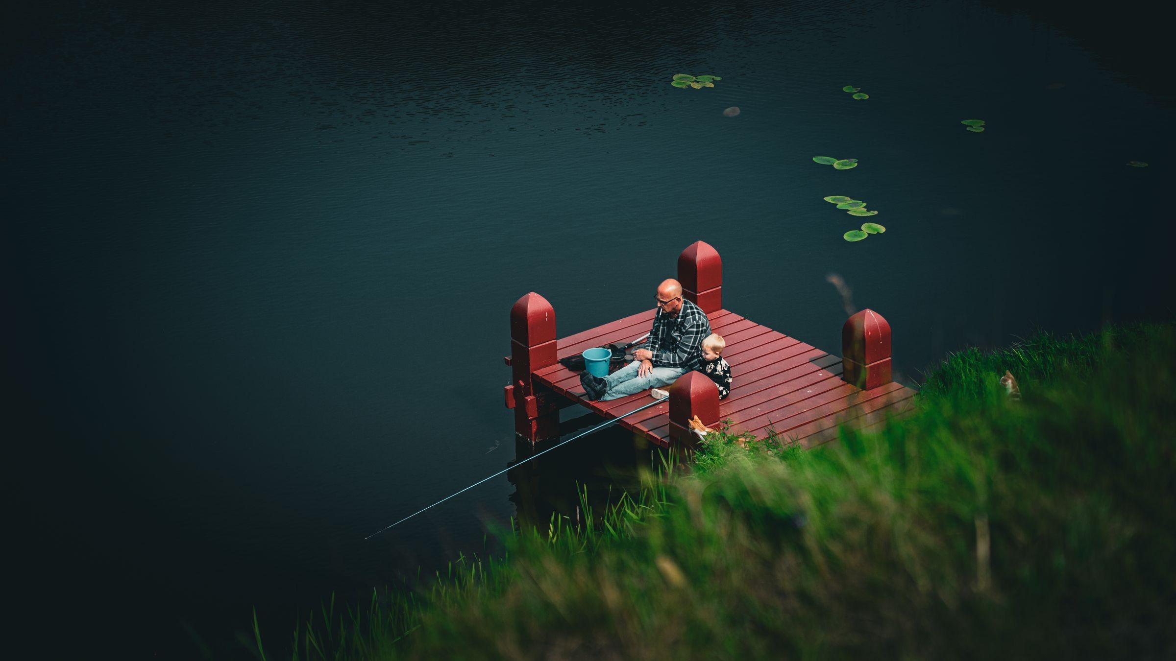 A parent and young child, seen from a distance, fish together from the end of a small red dock on a dark lake