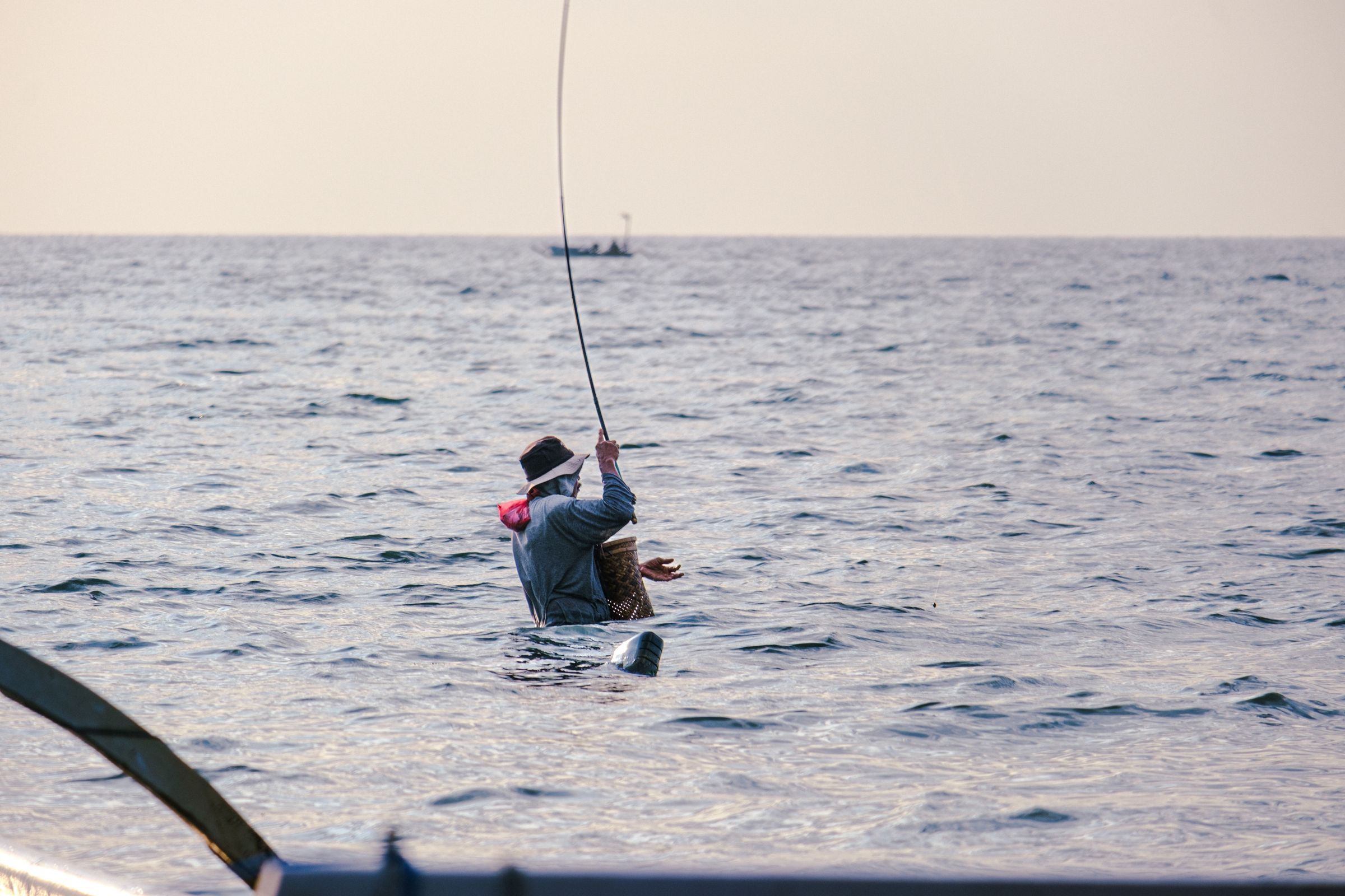An angler in a hat wades waist-deep in calm sea water with a bent rod