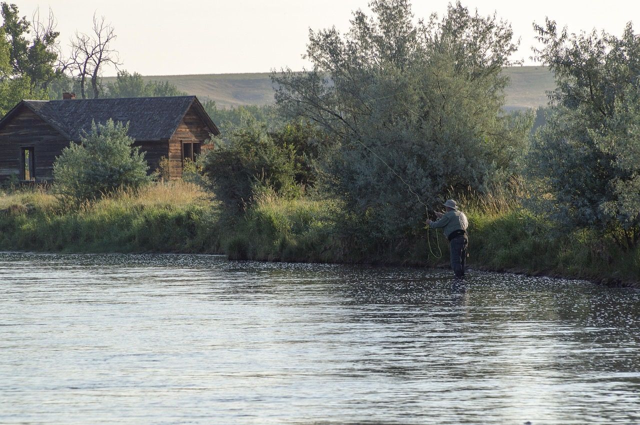 A fly angler wading a wide river in early light beside a weathered wooden cabin