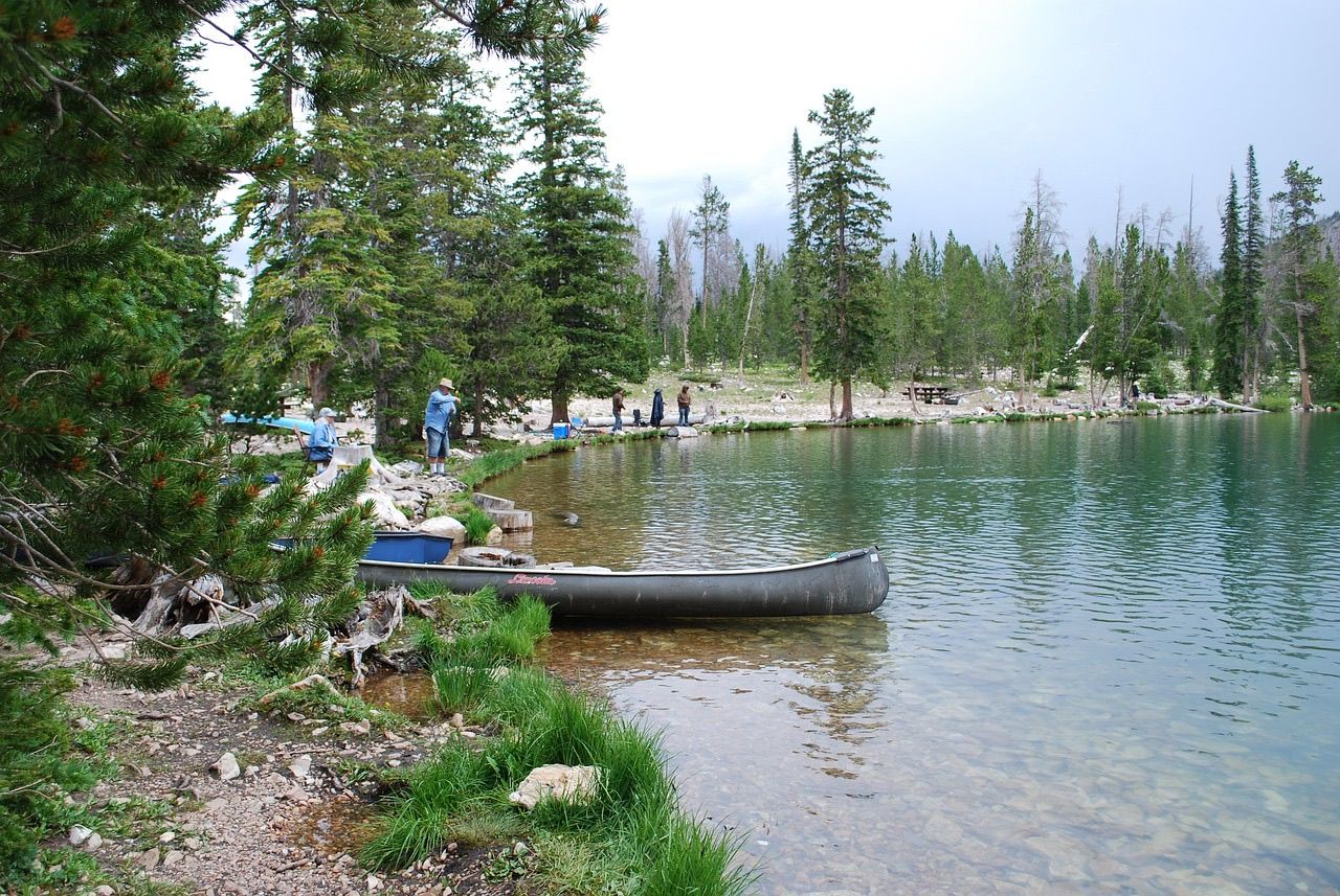 A canoe pulled up on the shore of a clear mountain lake with anglers fishing along the pine-lined bank