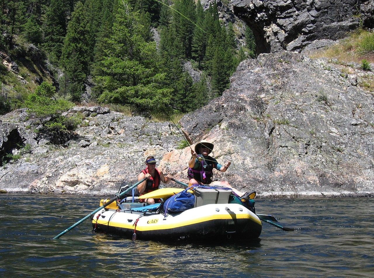 An oarsman rowing an inflatable raft while an angler fishes beside rocky cliffs on a forested river