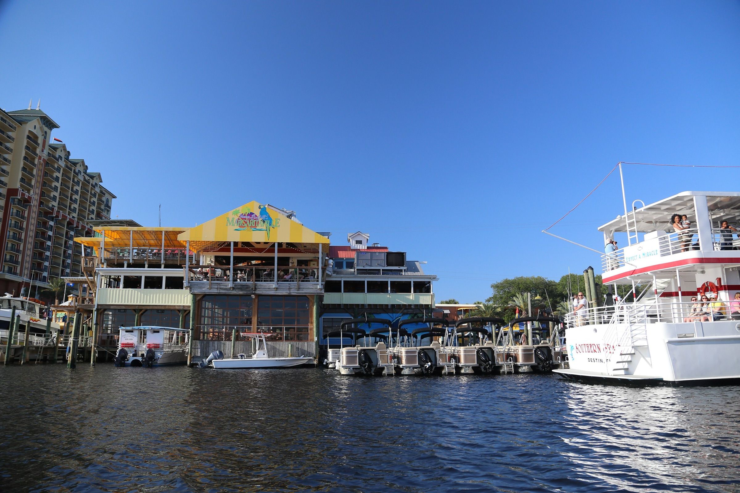 Waterfront restaurants, rental pontoon boats and a tour boat along the Destin harbor under a clear blue sky