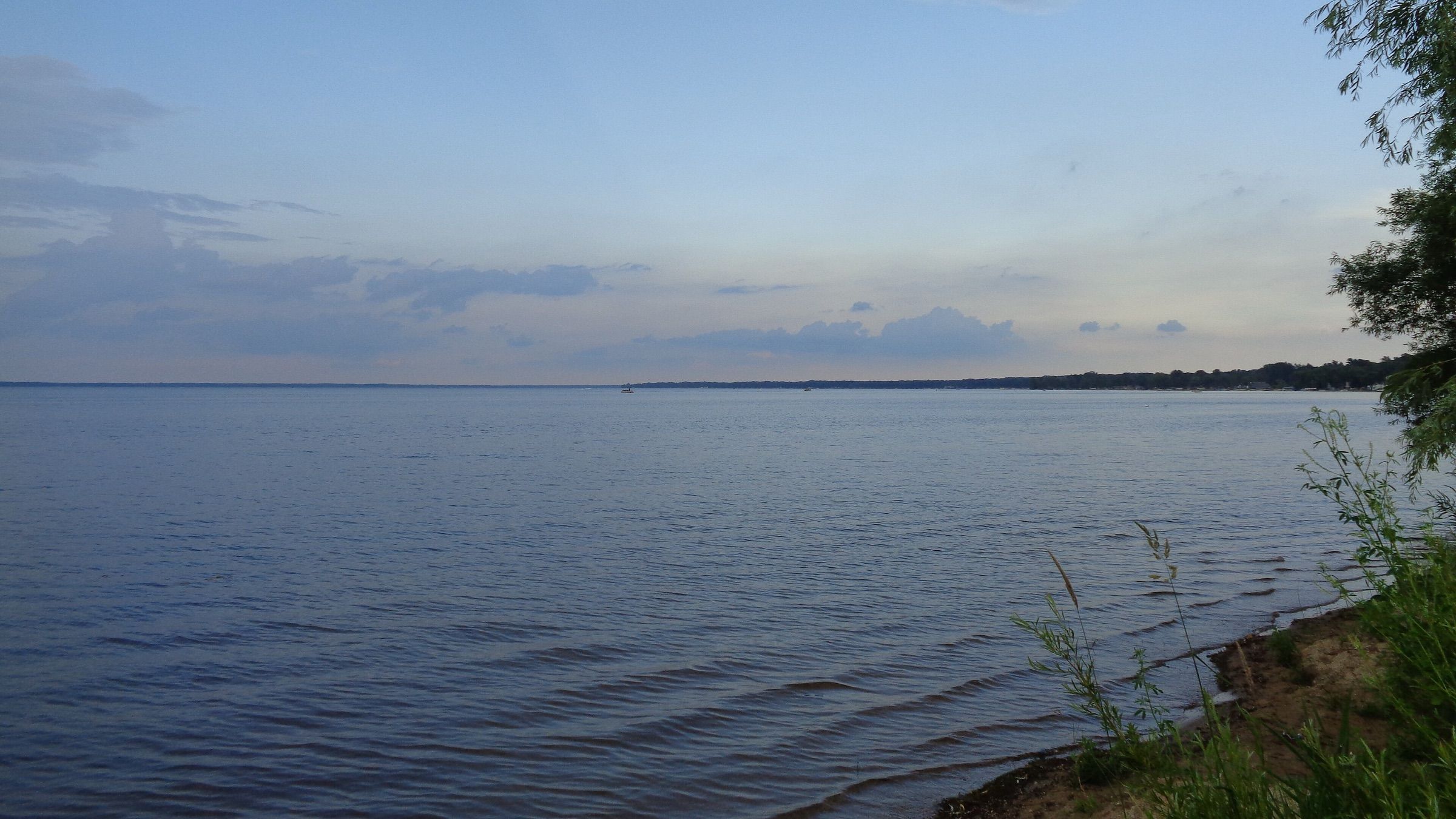 Calm open water of Houghton Lake at dusk with a wooded far shore and grasses in the foreground