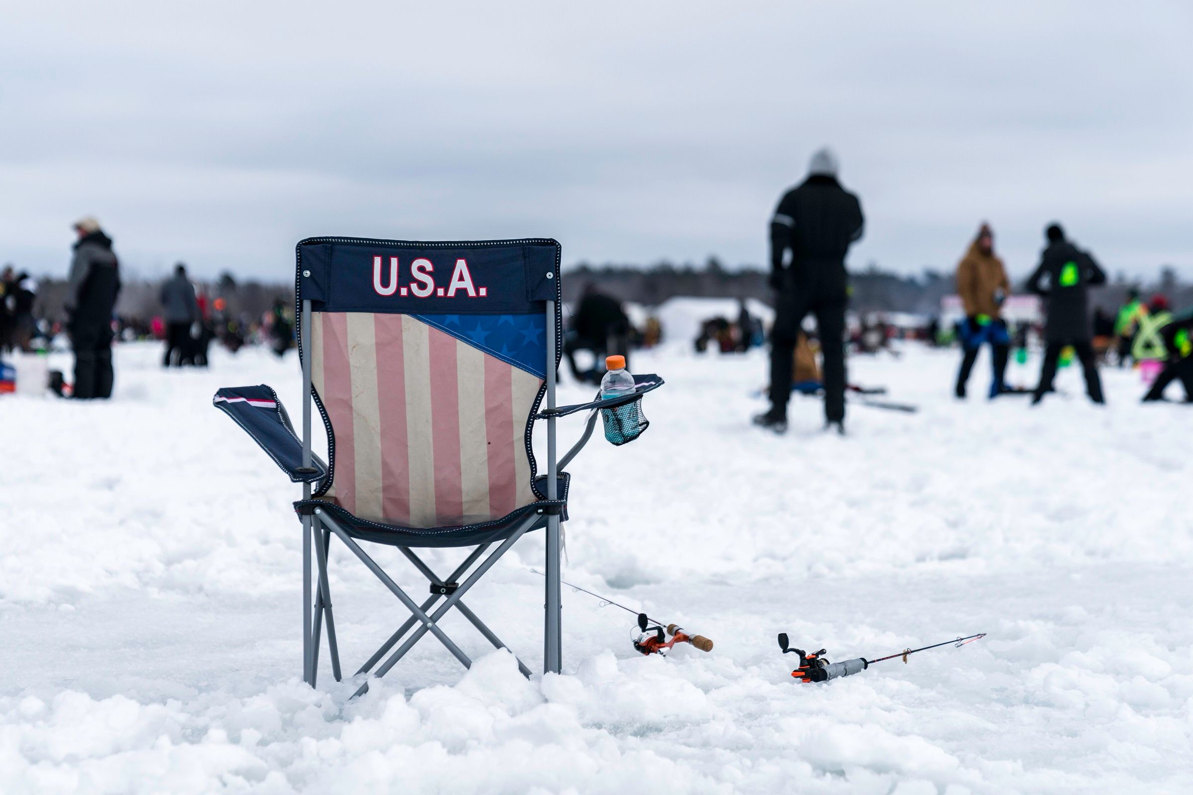 A folding camp chair and two short ice fishing rods resting on the snow on a frozen lake