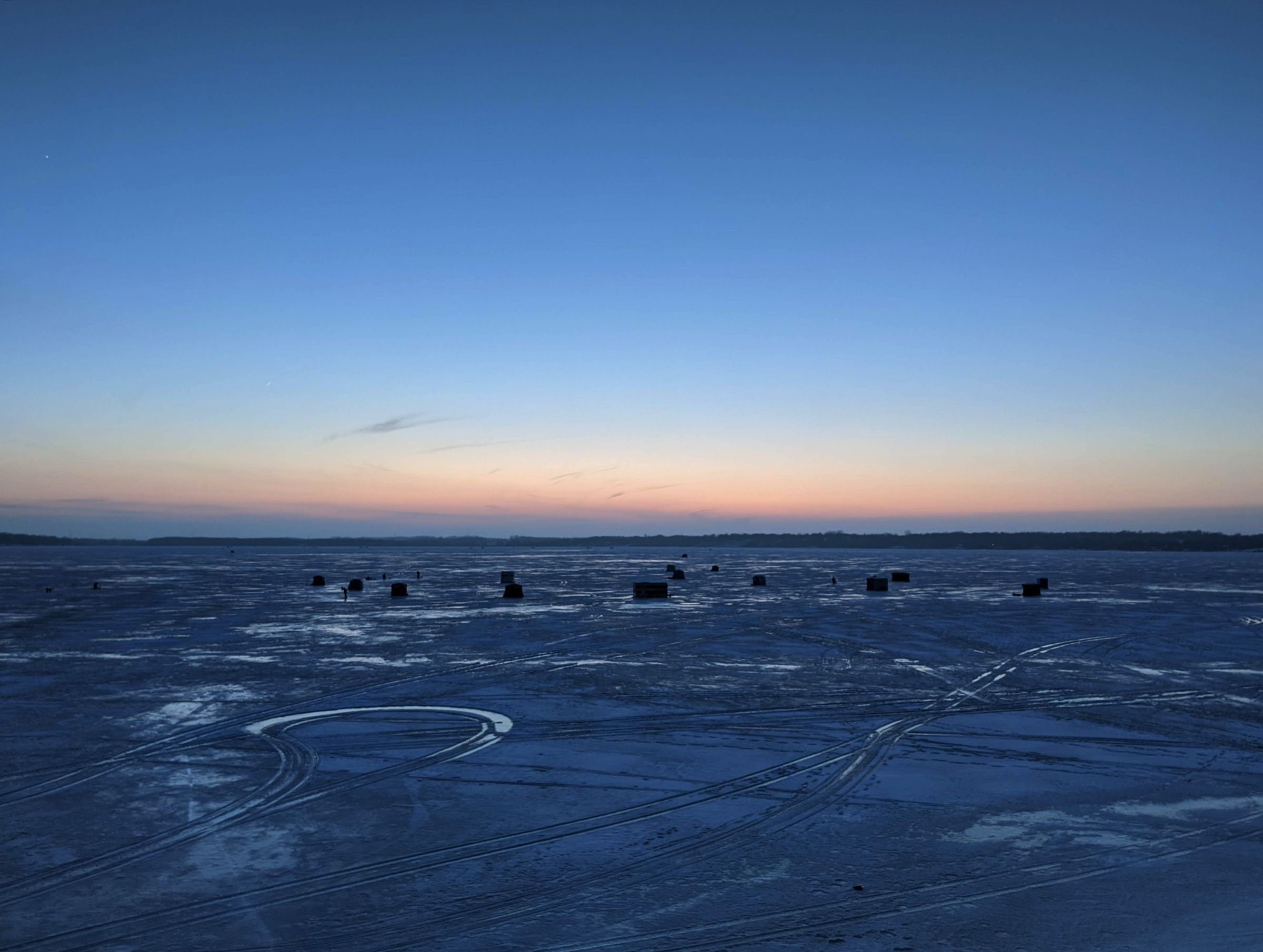Dark ice fishing shelters scattered across a frozen lake at dusk under a blue and orange sky