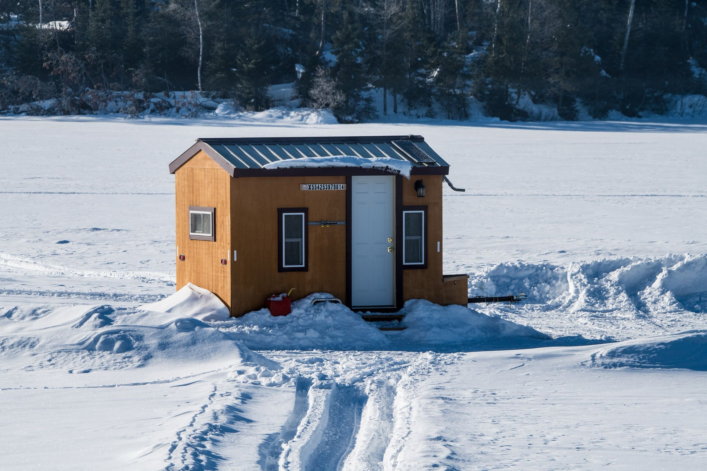 A wooden fish house with a metal roof sits on snow-covered lake ice, sled tracks leading to its door