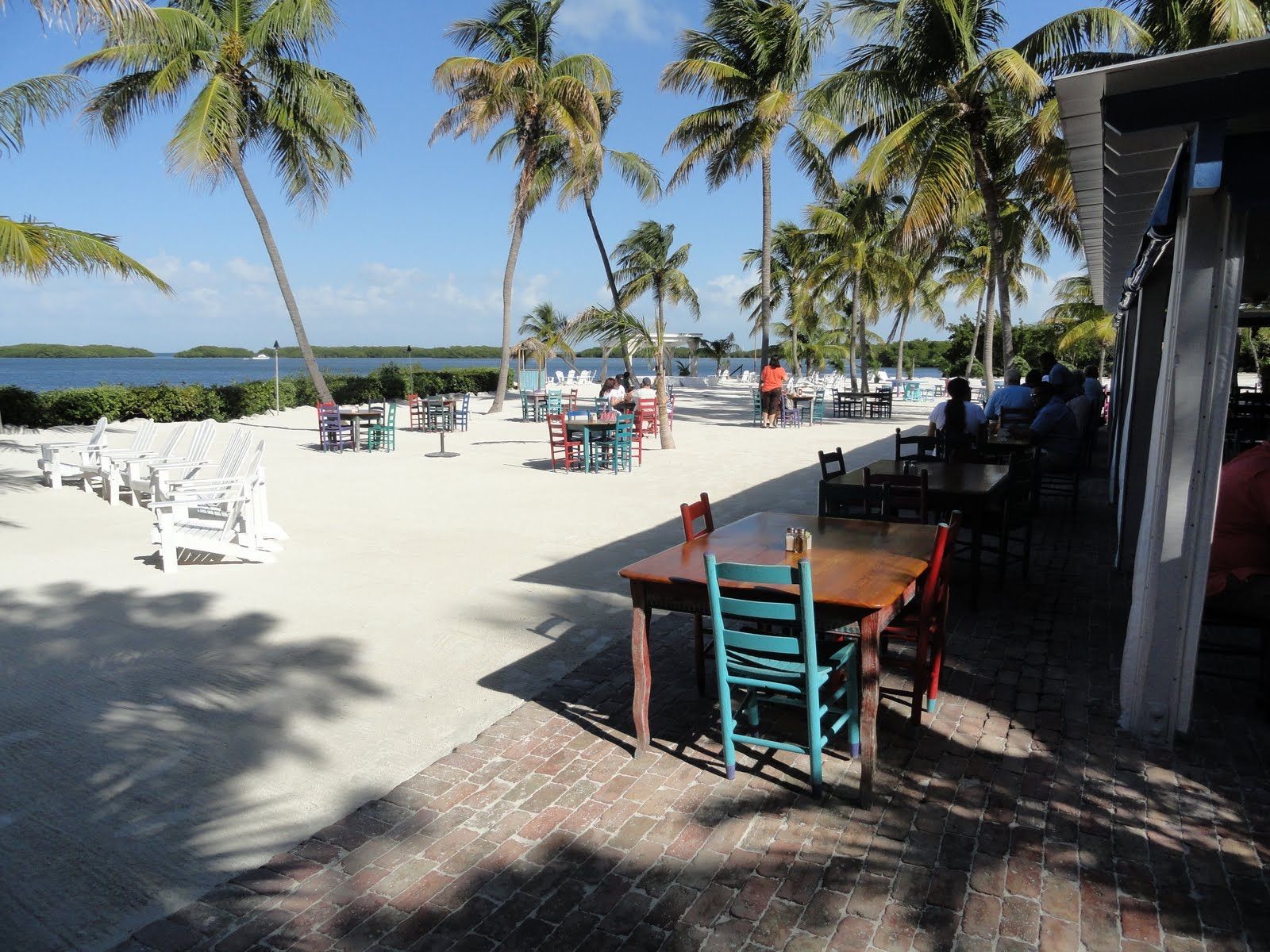 Colorful tables and Adirondack chairs on the sand under palm trees at the Morada Bay Cafe on the bay side of Islamorada