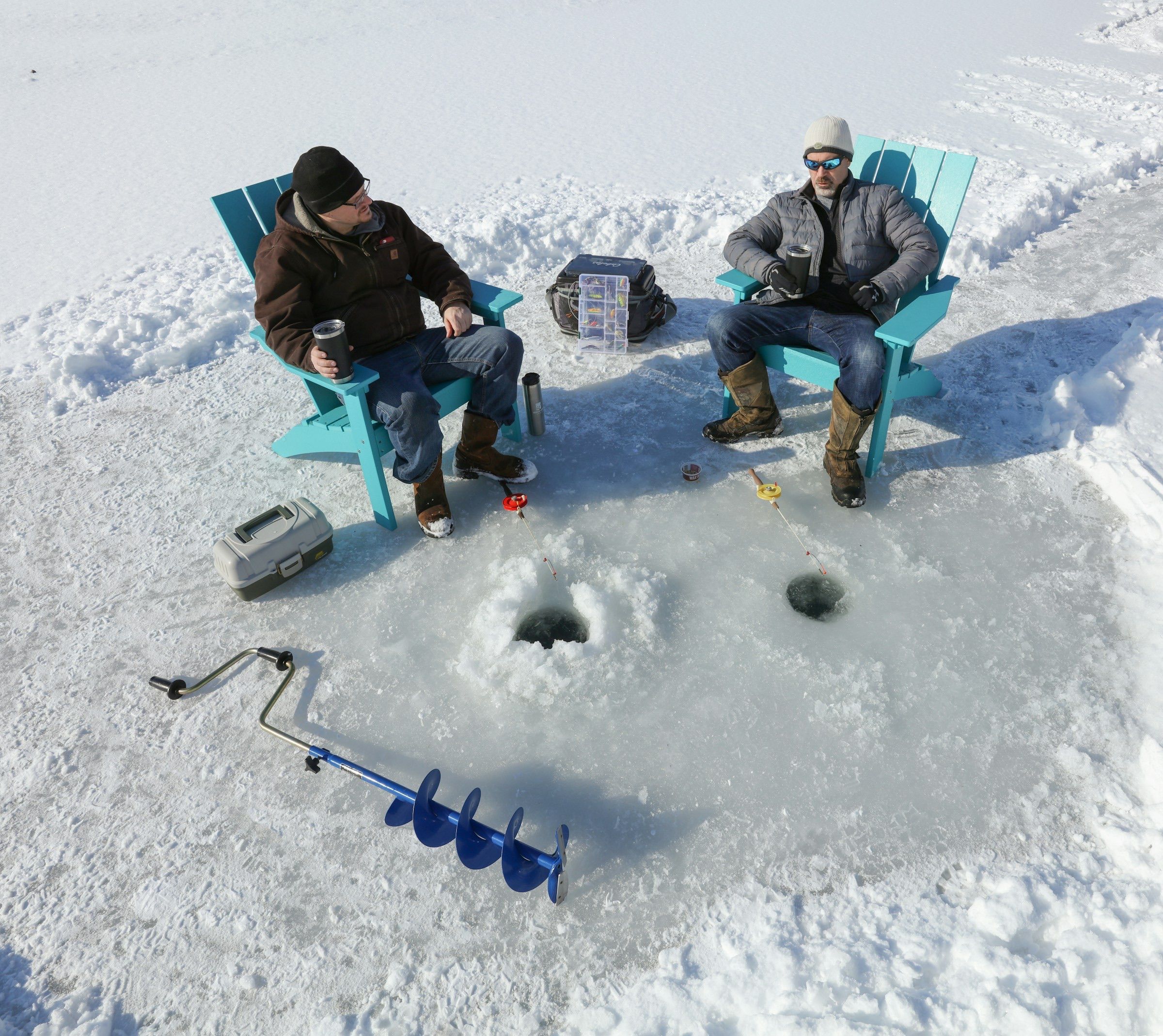 Two anglers in lawn chairs beside holes drilled through lake ice, with a hand auger on the snow
