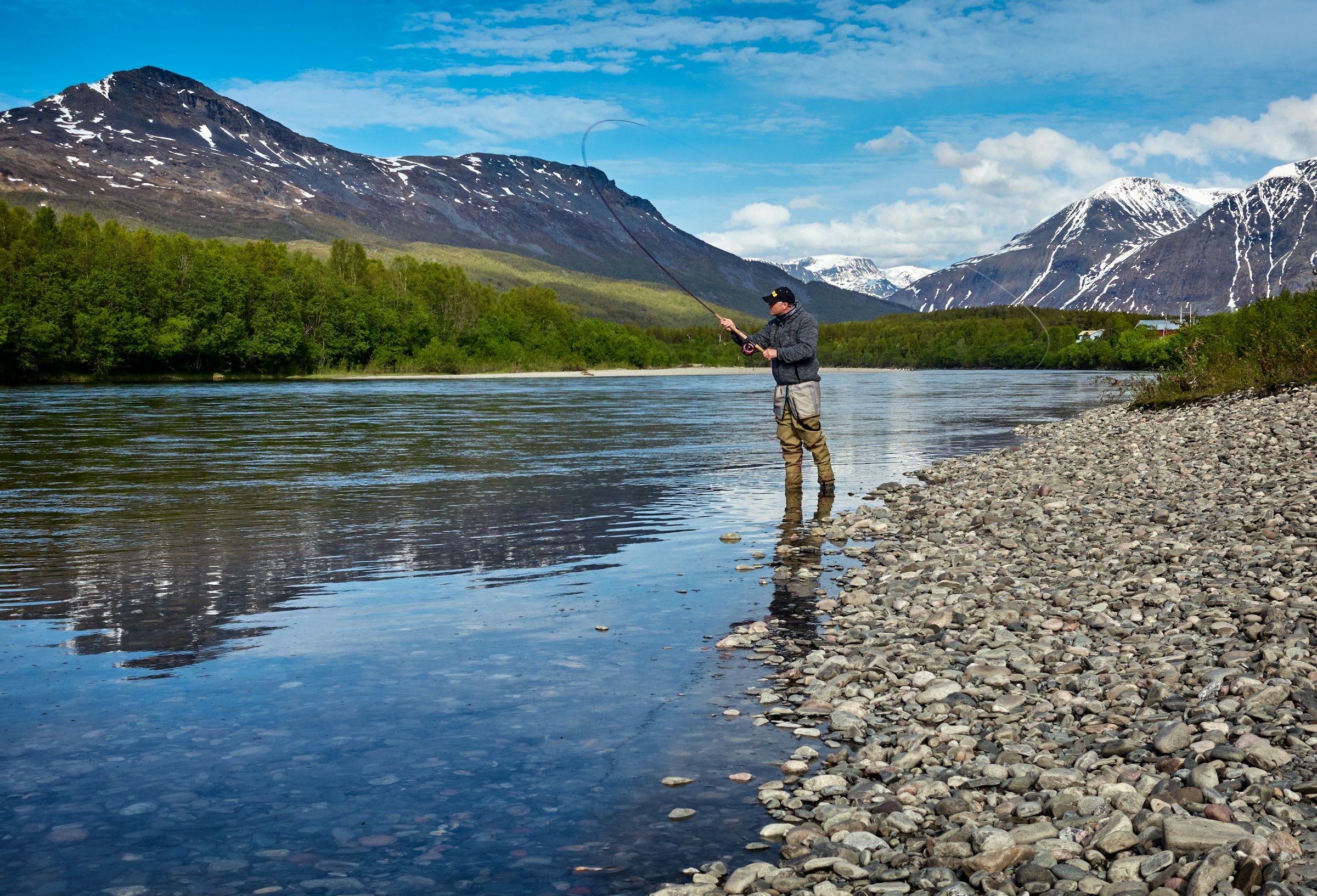Fly angler wading a clear river beneath green slopes and snow-streaked mountains