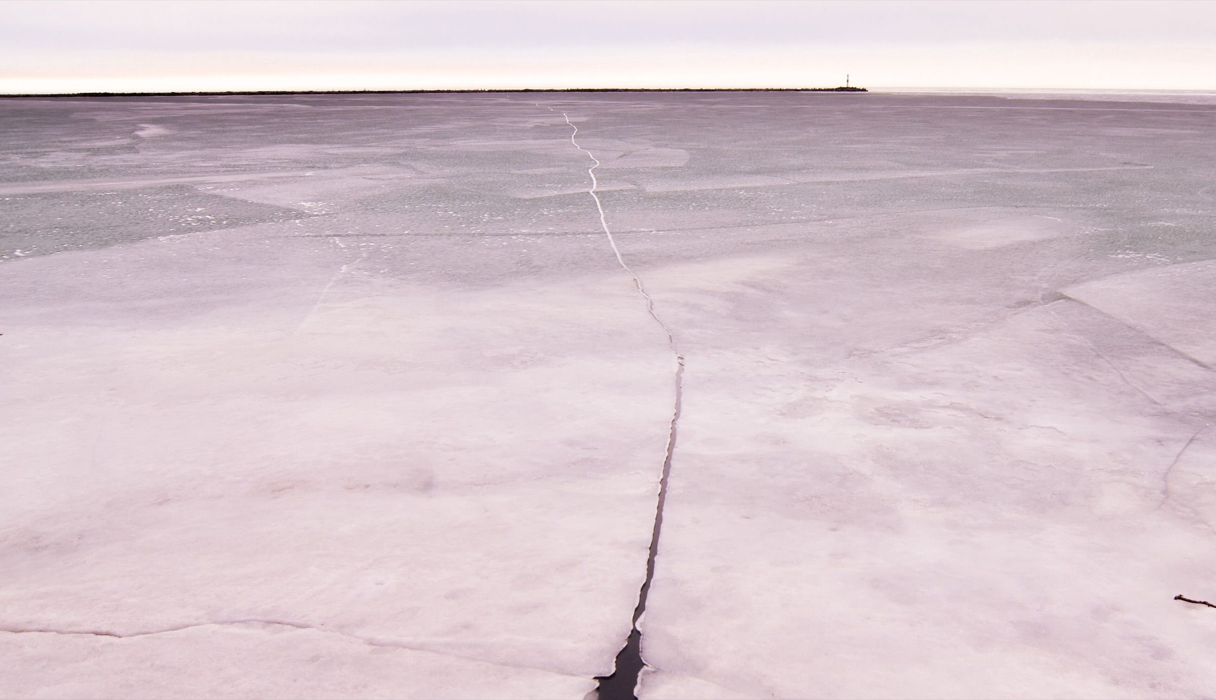 A long crack runs across the frozen surface of Lake Erie toward a distant breakwall light