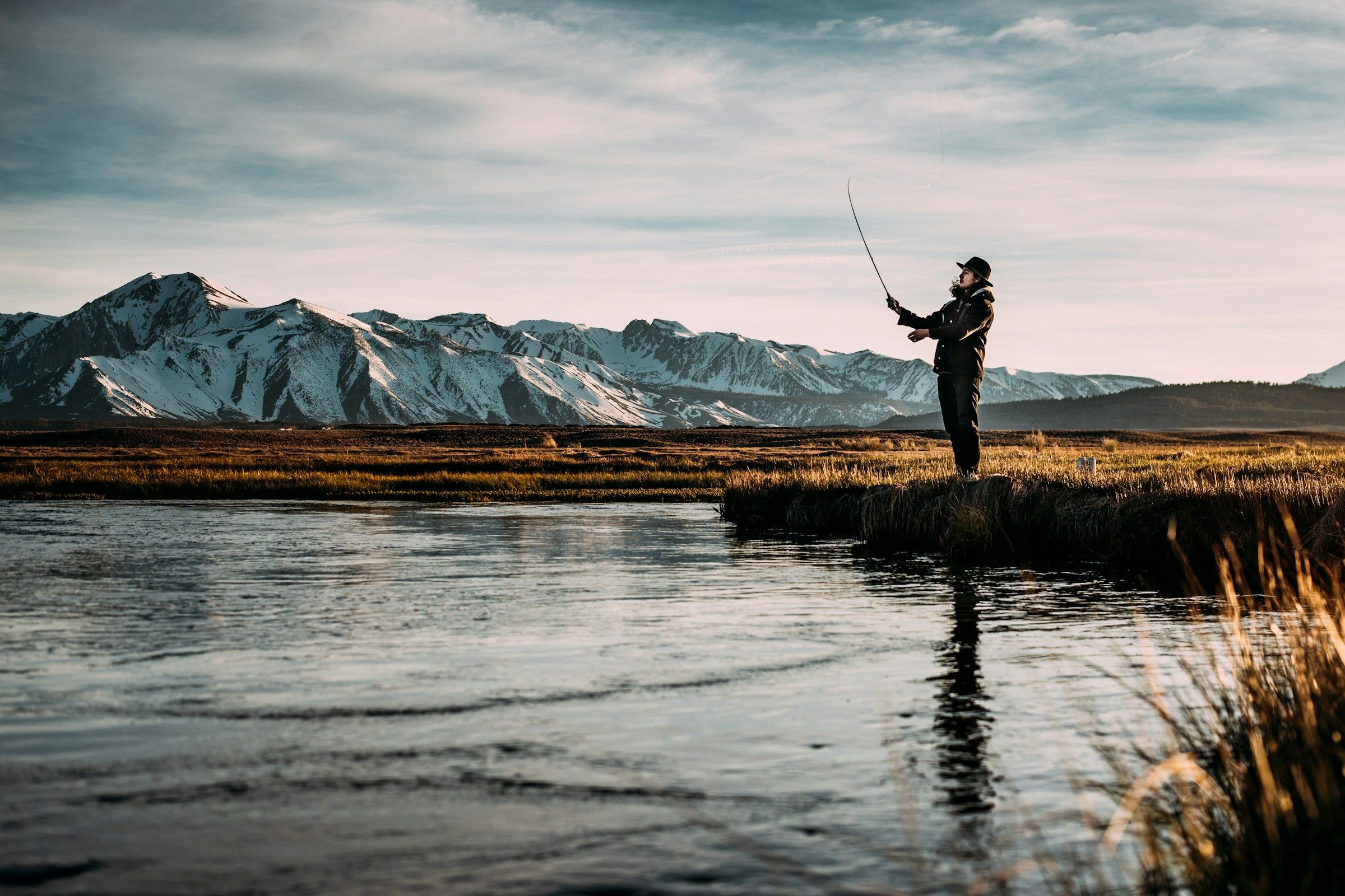 Fly angler casting on a calm river below snow-capped mountains