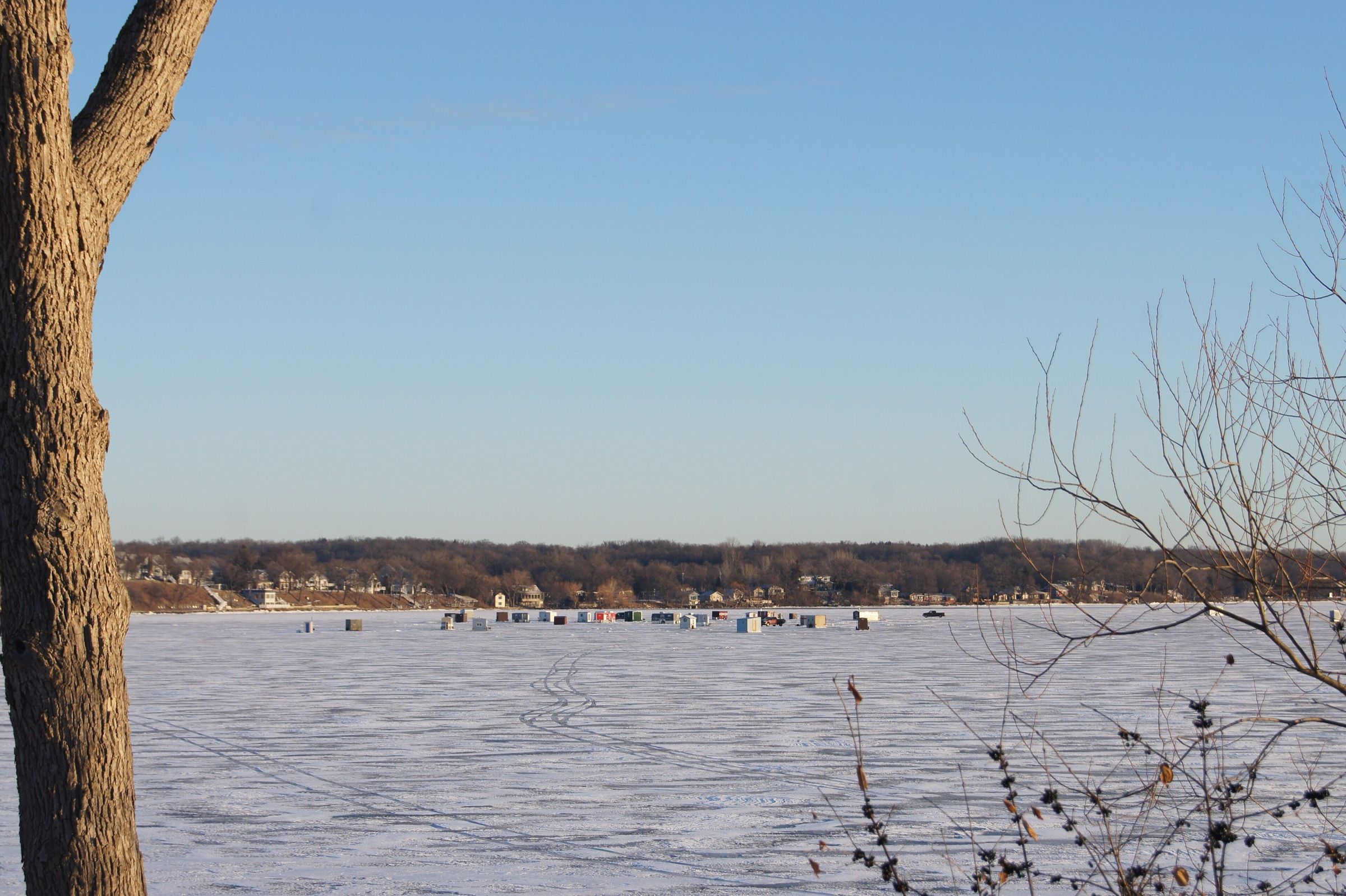 A cluster of fish houses on frozen Green Lake seen from the wooded shore on a clear winter day