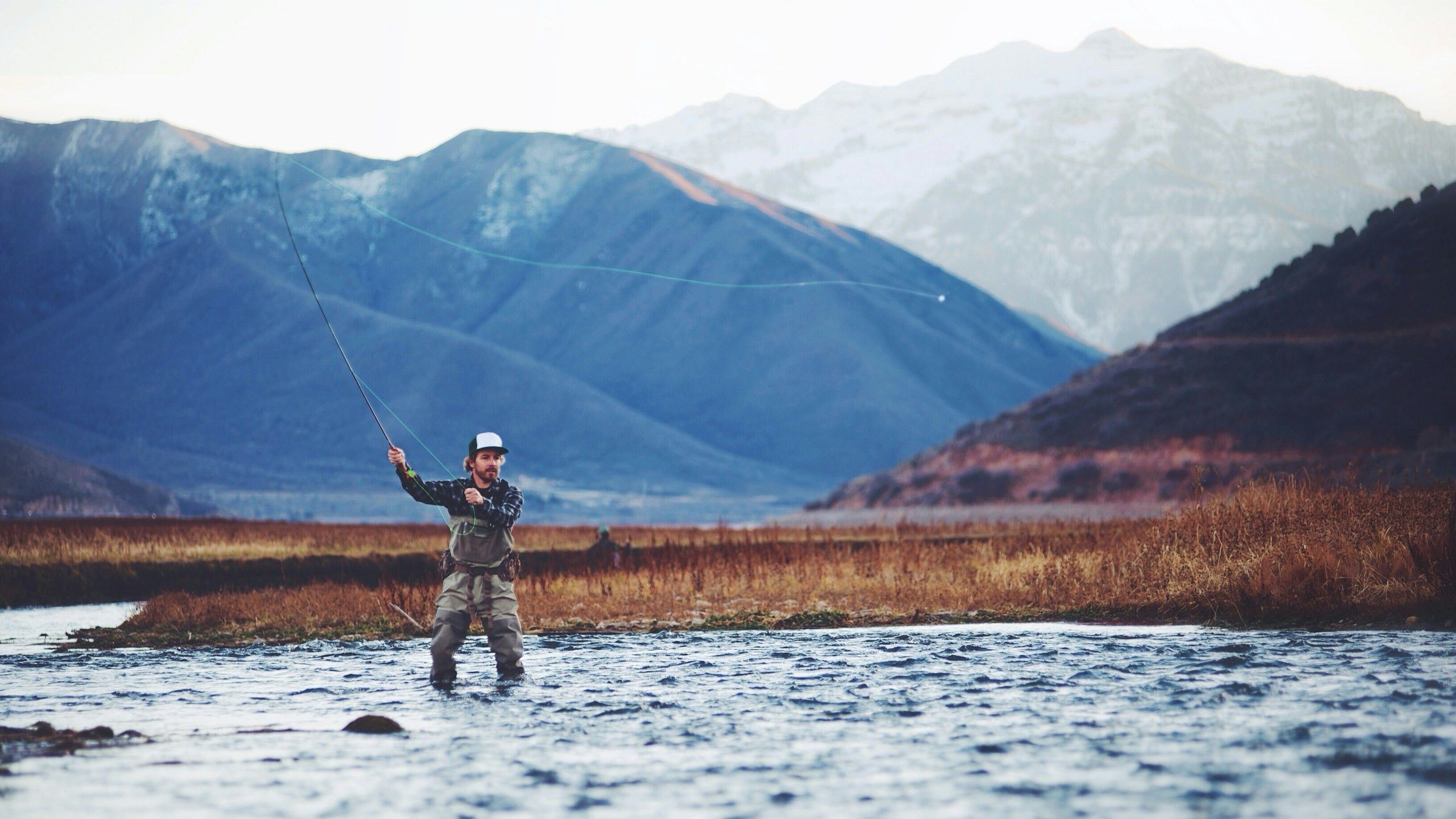 Fly angler casting in a cold river below snow-dusted mountains in late fall