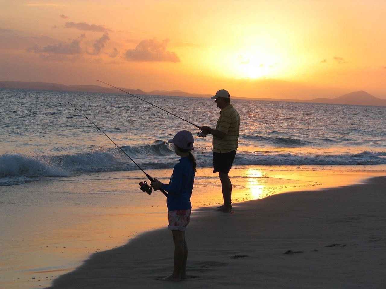 A parent and child silhouetted at sunset, each holding a fishing rod at the edge of a sandy beach