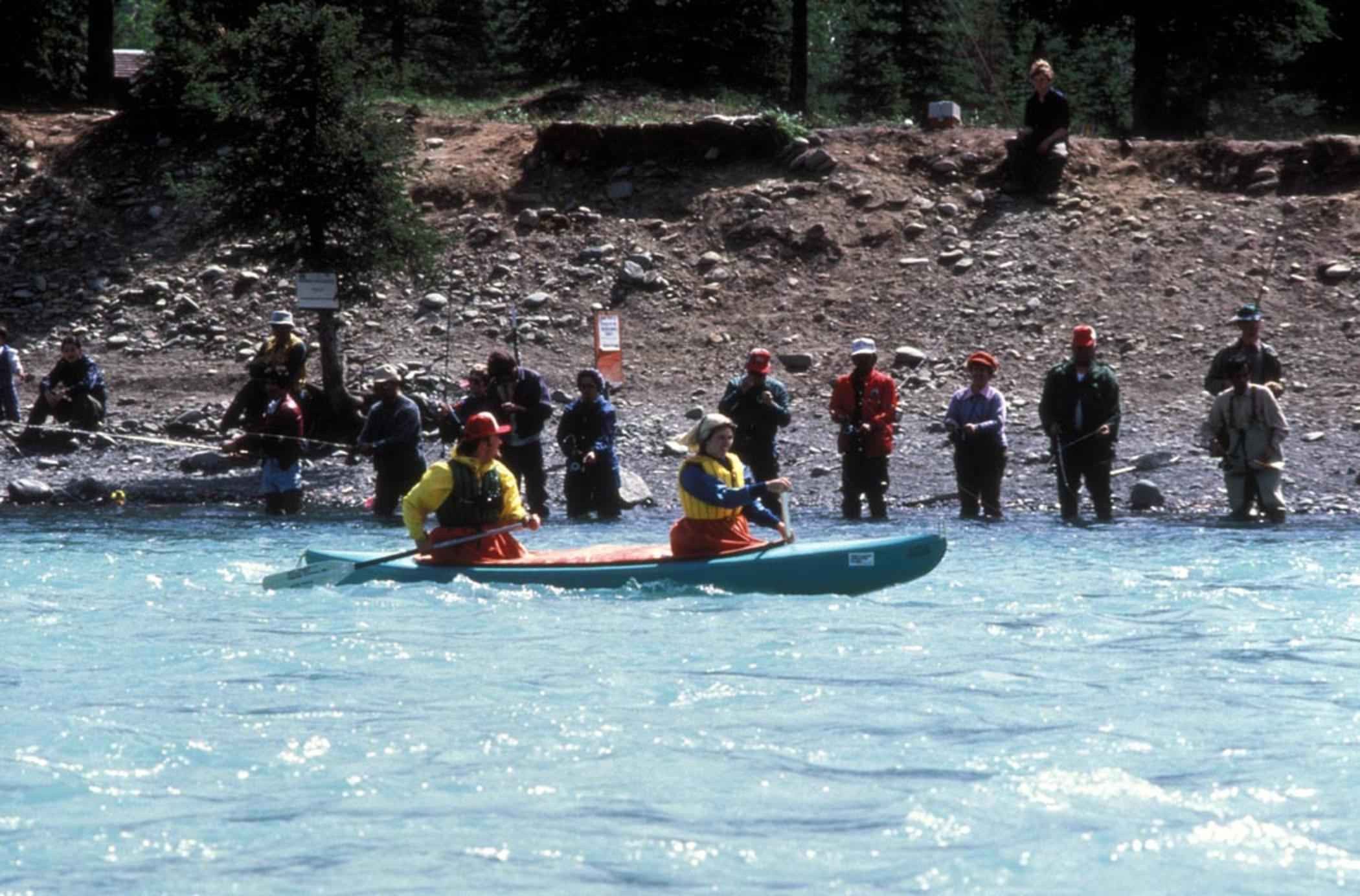 A line of anglers fishing shoulder to shoulder on the Kenai River as a canoe paddles past