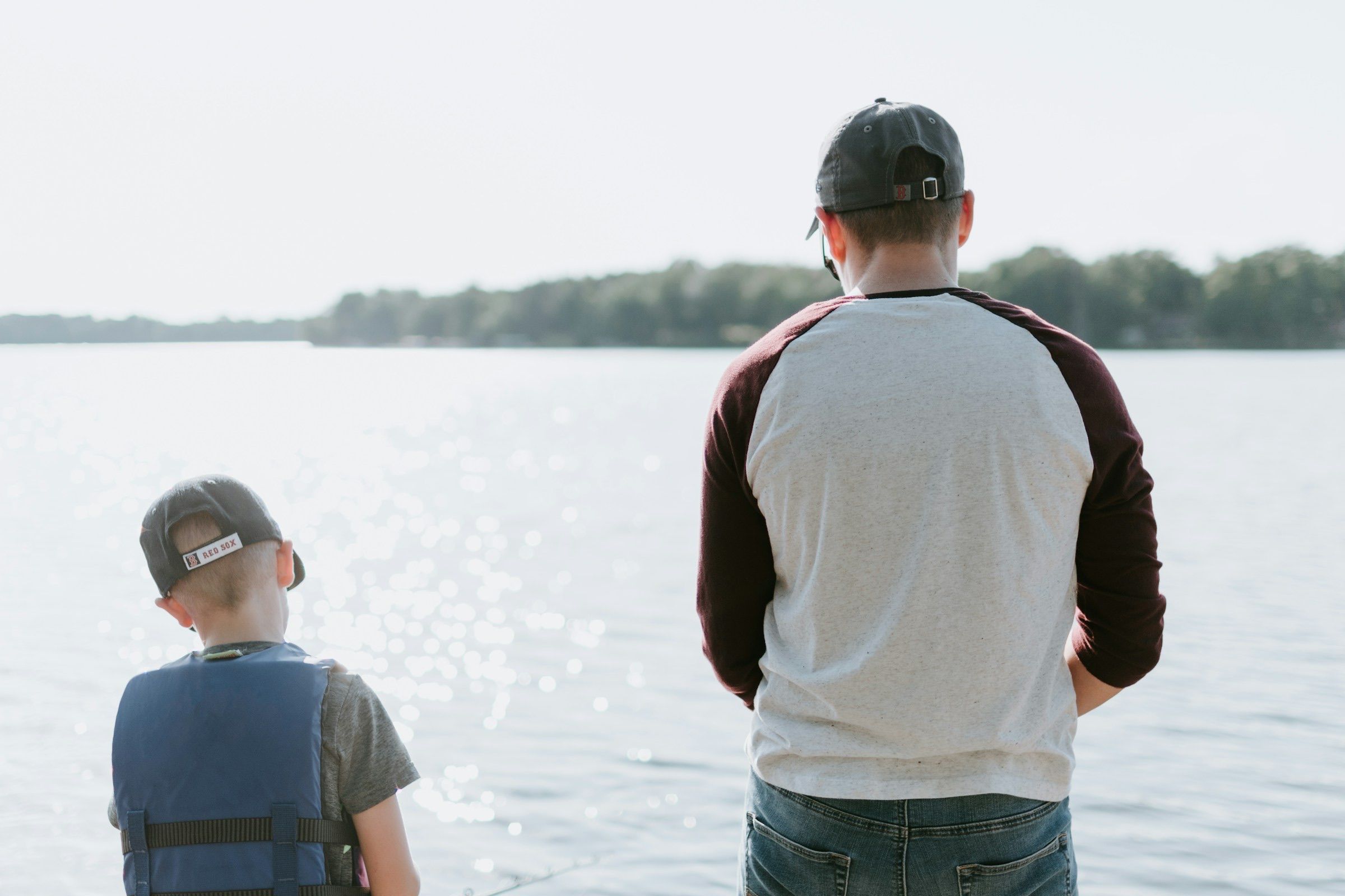 Adult and child in a life jacket seen from behind, fishing side by side on a lake