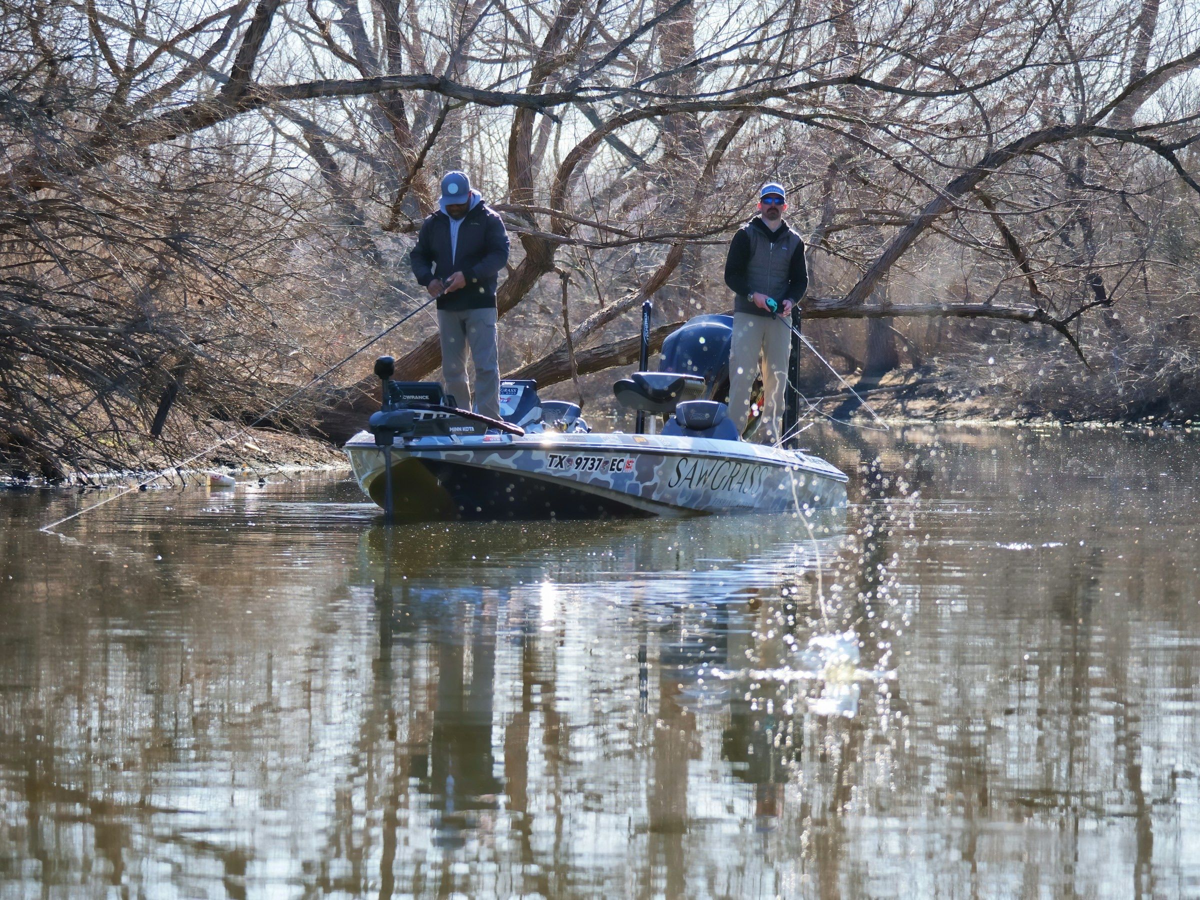 Two anglers casting from a bass boat along a flooded tree line in early spring