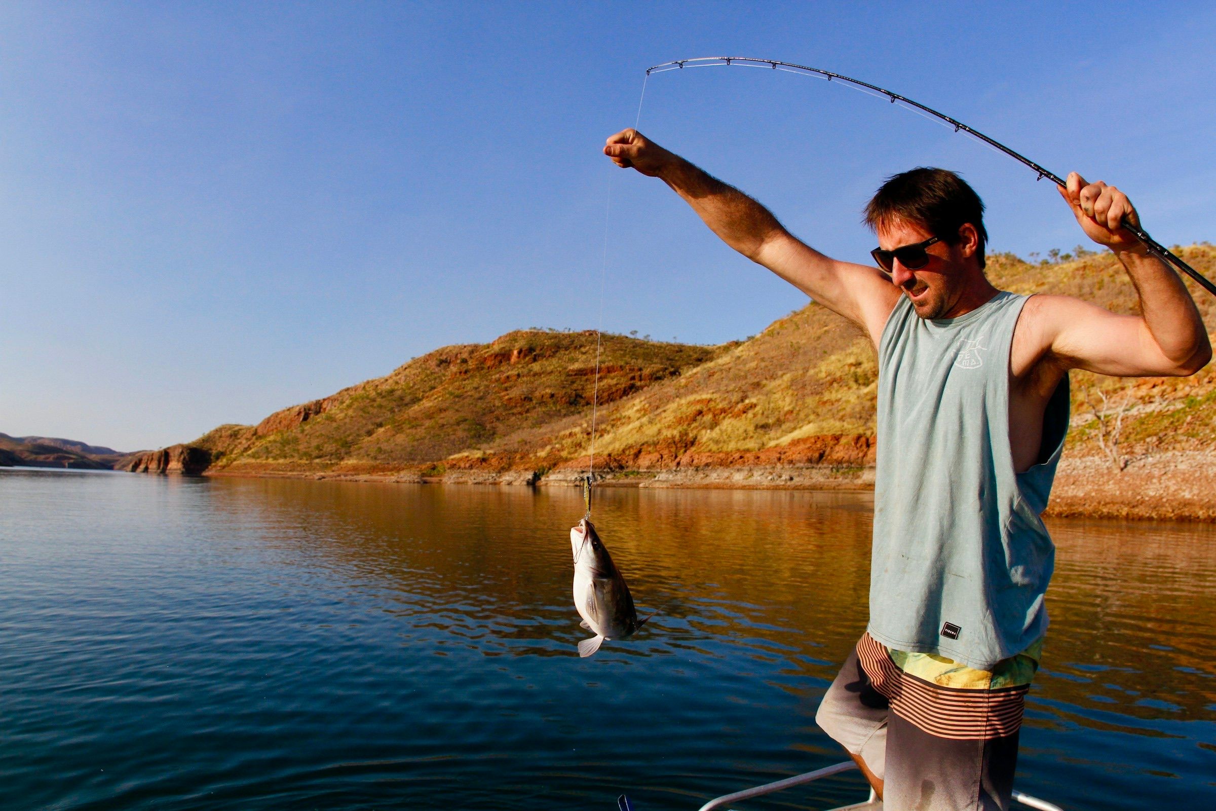 Angler in sunglasses bending a rod as he lifts a fish from a calm lake on a sunny day