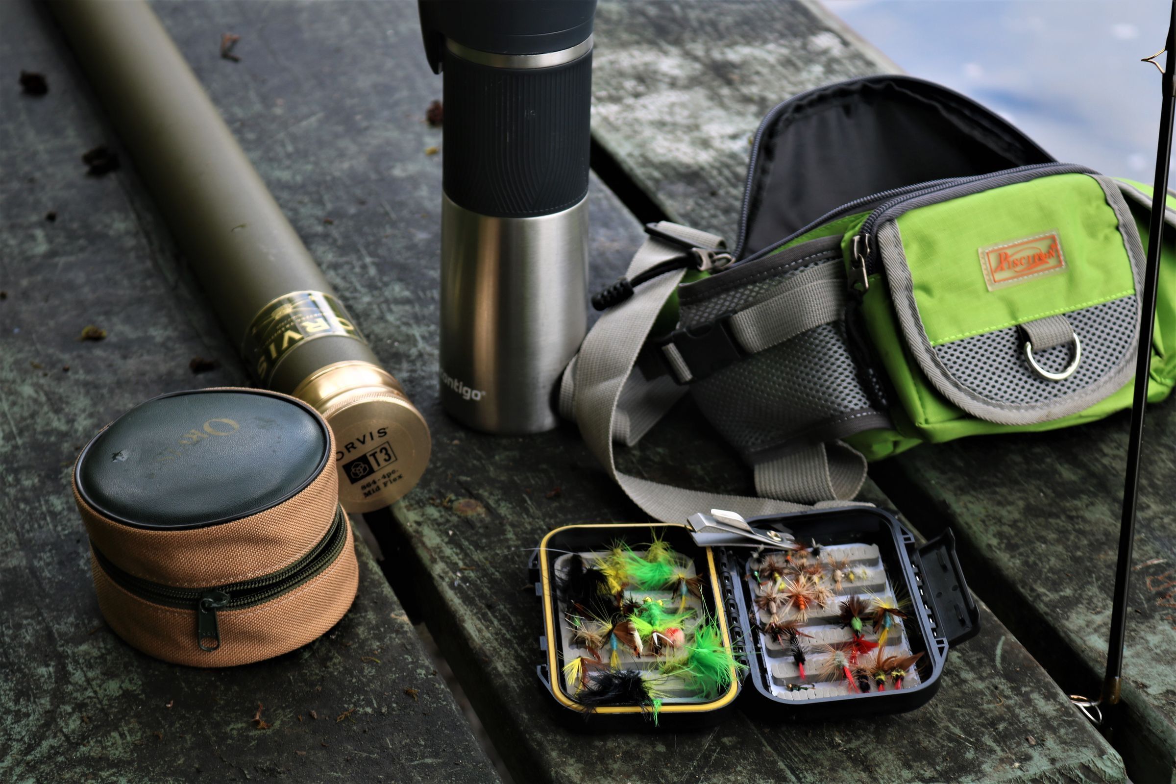 A fly rod tube, reel case, thermos, hip pack and box of flies set out on a wooden bench
