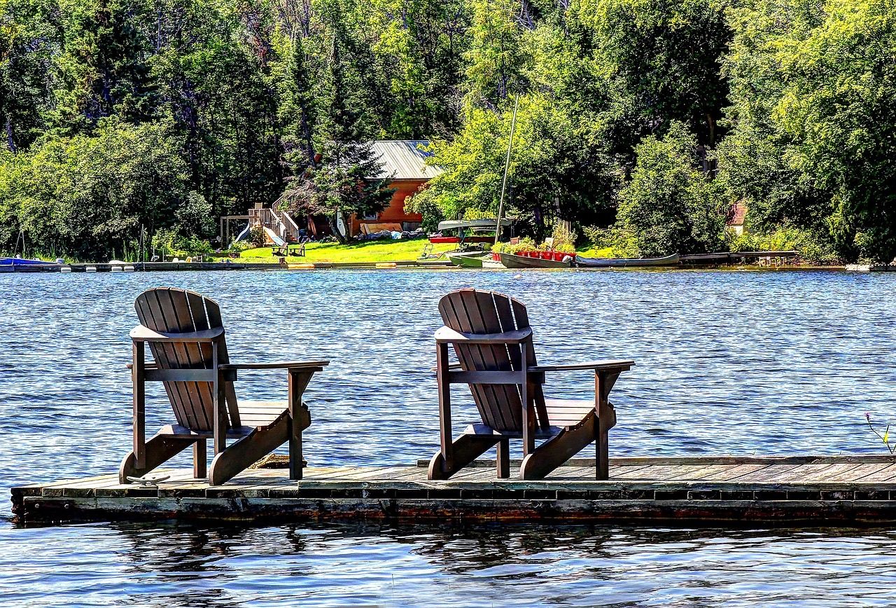 Two Adirondack chairs on a wooden dock looking across a lake to a cottage in the trees