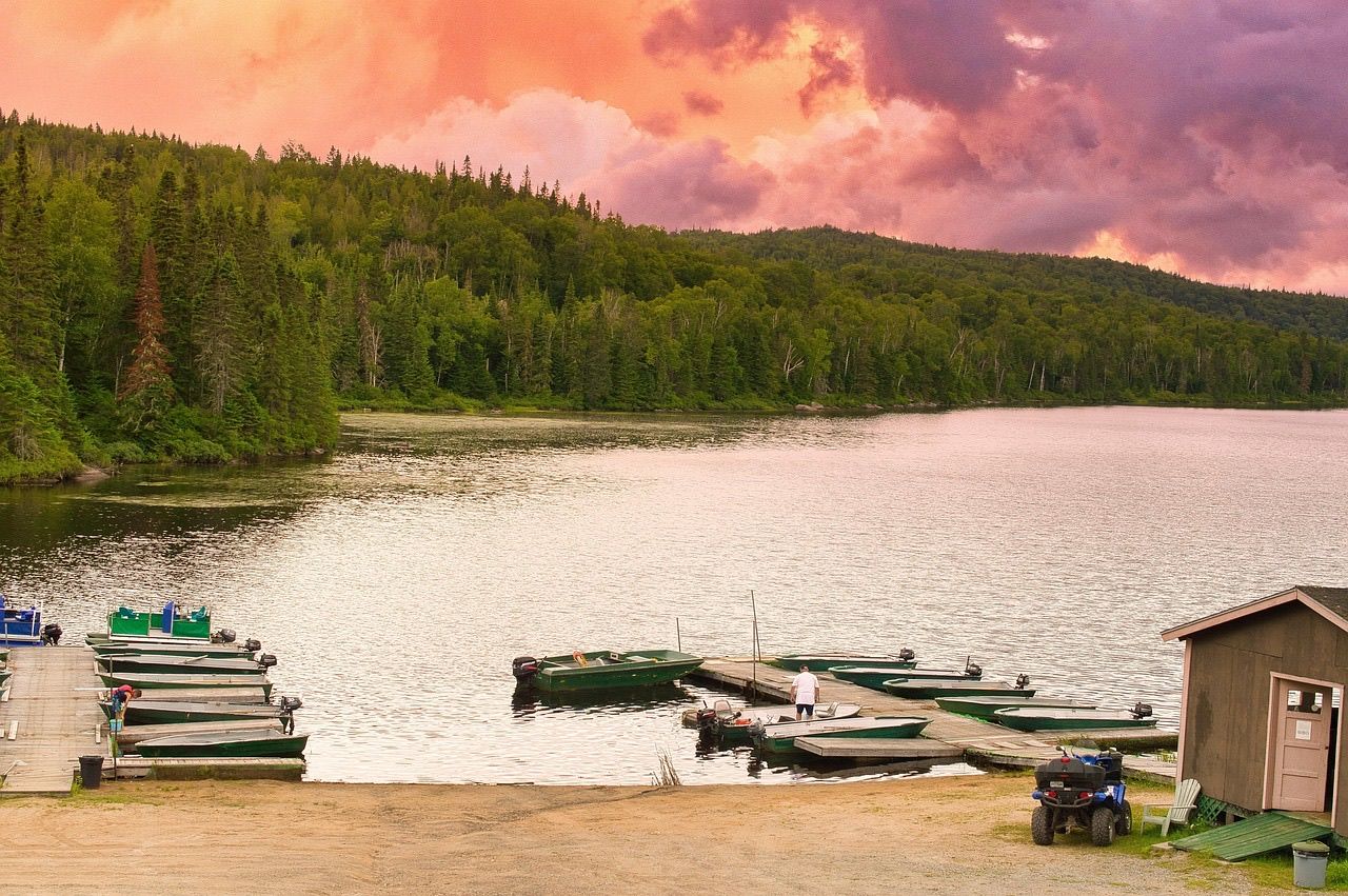 Small fishing boats tied along a dock below a forested lake shore under a pink evening sky